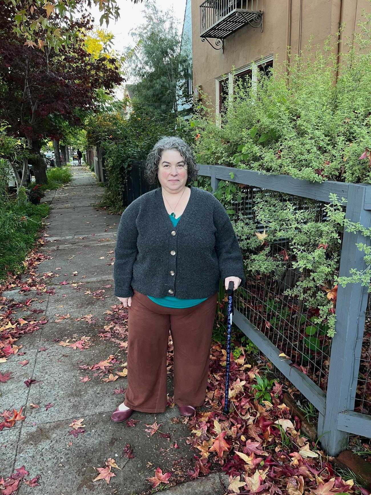 Rebecca Novick standing with her cane on a leafy sidewalk