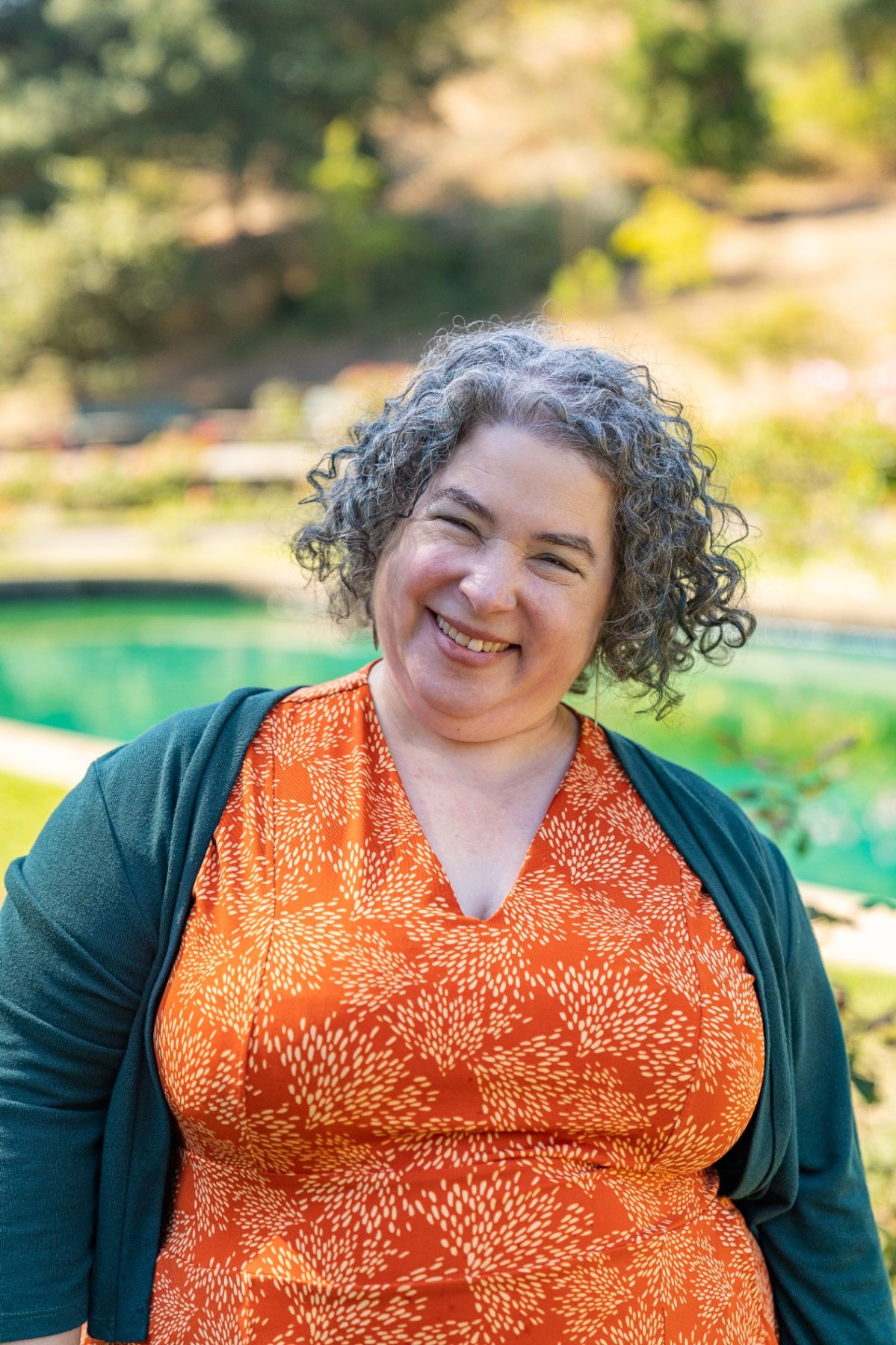 Rebecca Novick smiling in a garden, wearing an orange dress and teal cardigan